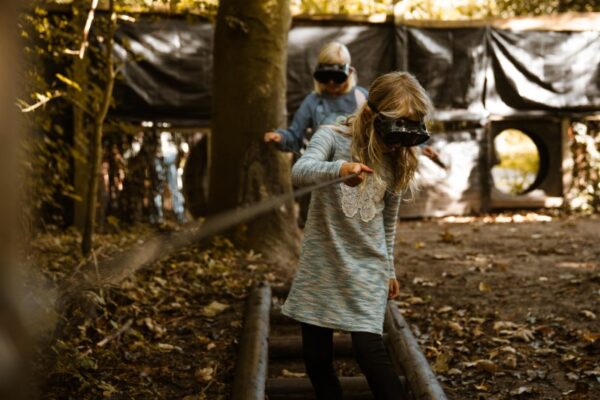 blinddoek-parcours-schoolreisje-teambuilding-outdoorpark-almere - Outdoorpark SEC Almere Twee geblinddoekte kinderen navigeren een boomstamparcours met een touw, een teambuildingactiviteit op schoolreisje bij Outdoorpark Almere.