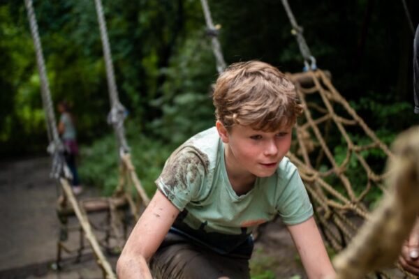 jongen-klimt-survival-hindernisbaan-modder-schoolreisje-almere - Outdoorpark SEC Almere Een jongen vol concentratie en modder klimt op een touwnet-hindernisbaan tijdens een survival schoolreisje in Almere
