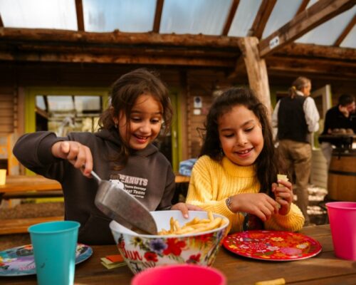 kinderen-eten-pauze-schoolreisje-outdoorpark-almere - Outdoorpark SEC Almere Twee blije kinderen eten frietjes tijdens een gezellige pauze op het buitenterras van Outdoorpark SEC Almere tijdens een schoolreisje.