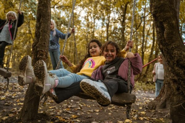 kinderen-schommelen-natuur-schoolreisje-outdoorpark-almere - Outdoorpark SEC Almere Twee blije kinderen schommelen op een houten schommel in een bosrijke omgeving tijdens een schoolreisje.