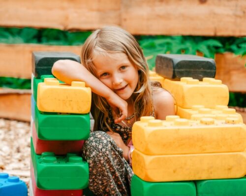 lachend-meisje-speelt-met-kleurrijke-blokken-outdoorpark-almere - Outdoorpark SEC Almere Lachend meisje met blonde haren spelend met grote, kleurrijke bouwblokken tijdens een schoolreisje of kinderactiviteit bij Outdoorpark SEC Almere, ideaal voor schooluitjes en teamactiviteiten.