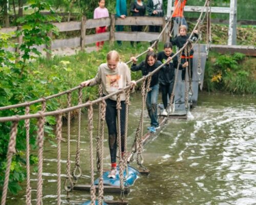 schoolreisje-kinderen-waterhindernisbaan-survival-outdoorpark-almere - Outdoorpark SEC Almere Groep leerlingen balanceren over een waterhindernisbaan met touwen en tonnen, begeleid door instructeurs, tijdens een survival schoolreisje bij Outdoorpark SEC Almere in de natuur.