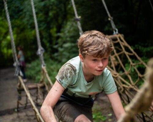 schoolreisje-survival-hindernisbaan-jongen-almere - Outdoorpark SEC Almere Jonge jongen bedwingt modderige hindernisbaan tijdens survival schoolreisje bij Outdoorpark SEC Almere, avontuurlijk uitje voor kinderen, touwbrug klimmen.