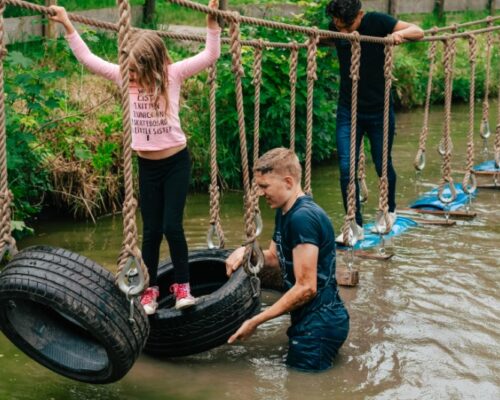 schoolreisje-survival-hindernisbaan-outdoorpark-almere - Outdoorpark SEC Almere Leerlingen en instructeur navigeren een survival hindernisbaan over water met touwen en banden tijdens een actief schoolreisje bij Outdoorpark SEC Almere, focus op samenwerking en avontuur.